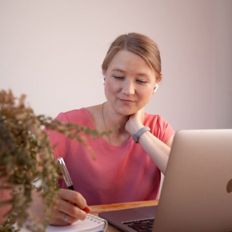 Junge Frau mit Kopfhörern, die am Laptop arbeitet und Notizen macht. Pflanze im Vordergrund.