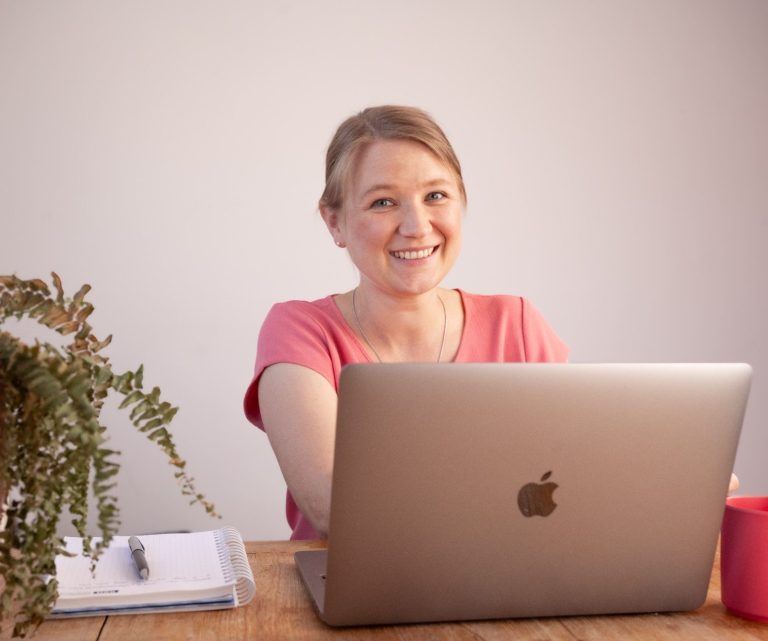 Lächelnde Frau mit Laptop, Notizblock und Pflanze auf einem Holztisch.