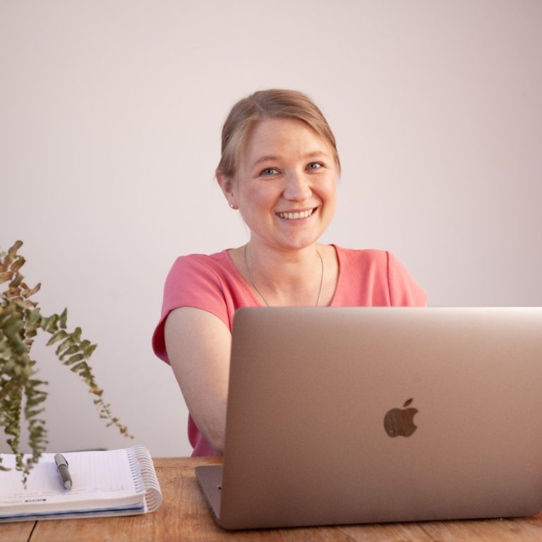 Eine lächelnde Frau sitzt vor einem Laptop auf einem Holztisch. Pflanzen sind im Hintergrund.