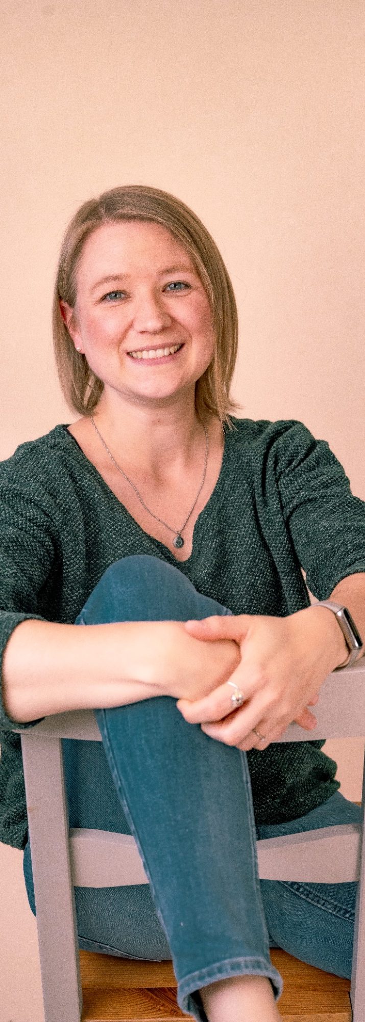 Eine lächelnde Frau sitzt entspannt auf einem Stuhl, mit blonden Haaren und grünem Oberteil.