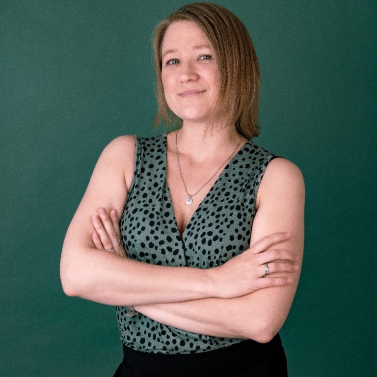 Frau mit kurzen Haaren, verschränkt die Arme, trägt ein gepunktetes Top vor grünem Hintergrund.